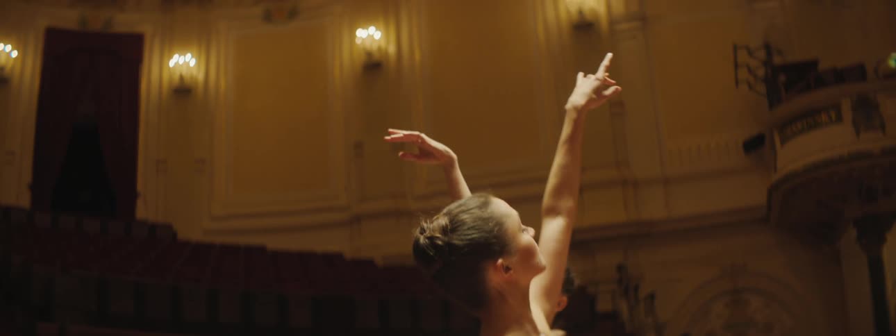 Two female ballerinas are dancing inside a dim theater
