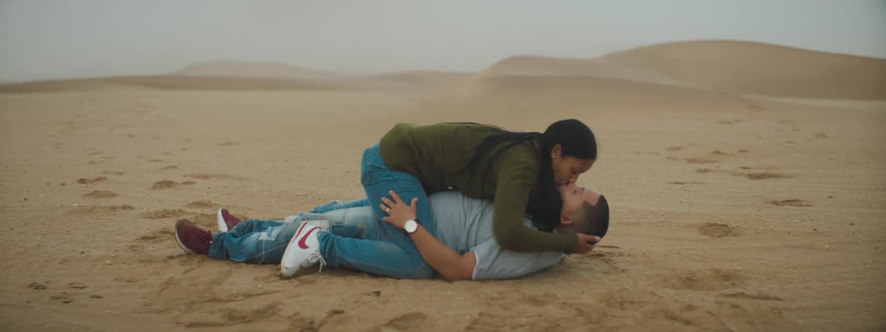 A man laying in the desert sand with a woman on top of him kissing him with the sky and clouds in the background