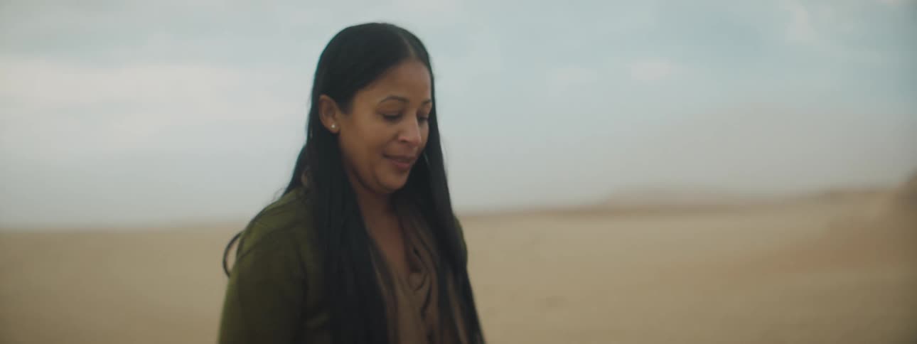 Brunette woman with a sweatshirt and jeans sits on man lying on the sand in the desert