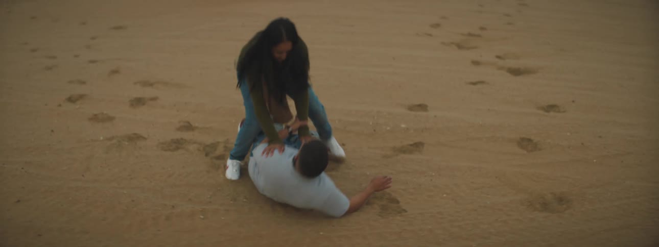 A man falling onto the desert sand with a woman sitting on top of him with the sky and clouds in the background