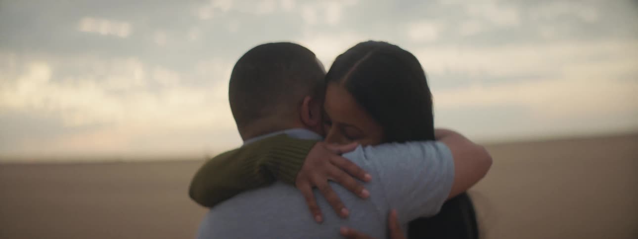 Man with a blue polo shirt and woman with long straight hair lovingly cuddle each other on the plain at dusk