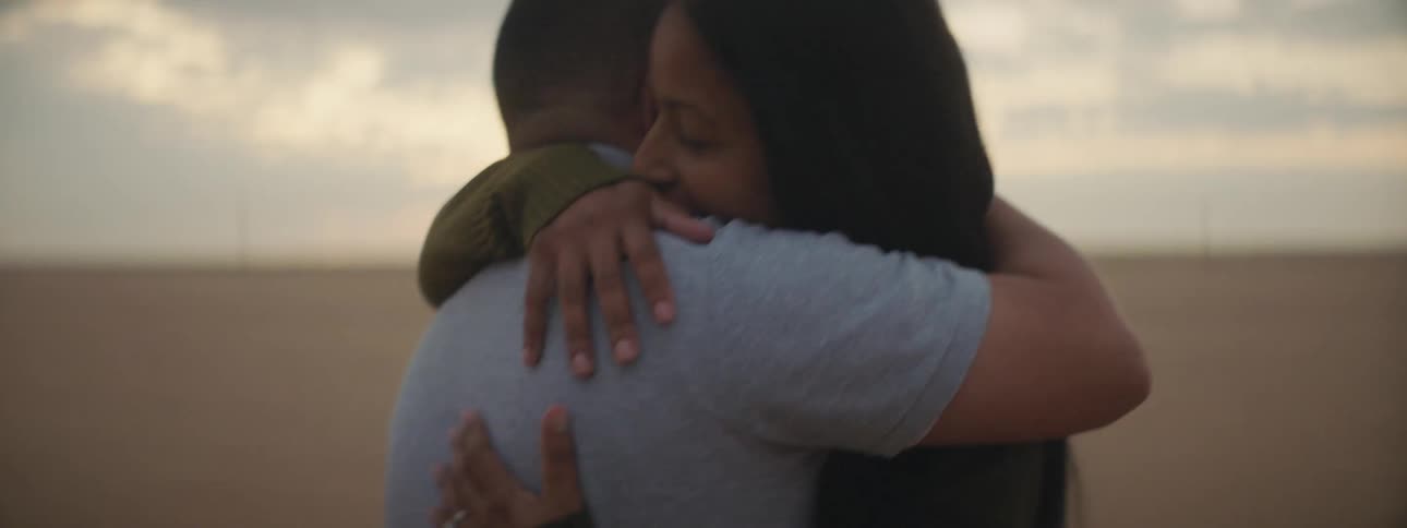 Man with a blue polo shirt and woman with long straight hair cuddle each other on the plain at dusk