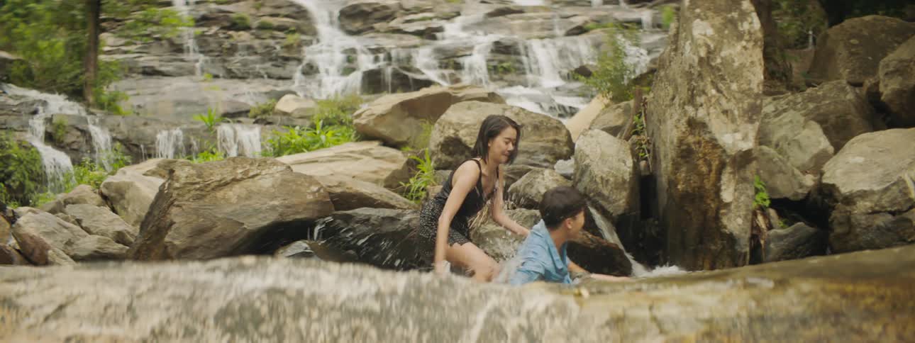 A boy and girl splash each other with water from a waterfall coming from large rocks