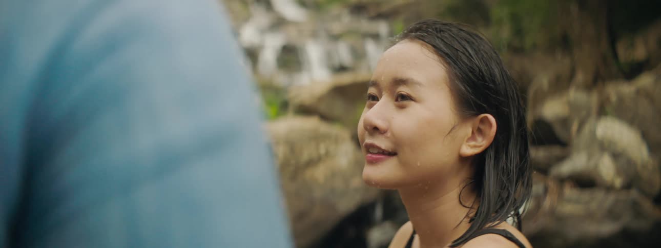 A woman wet with water with a waterfall a cliff and plants in the background