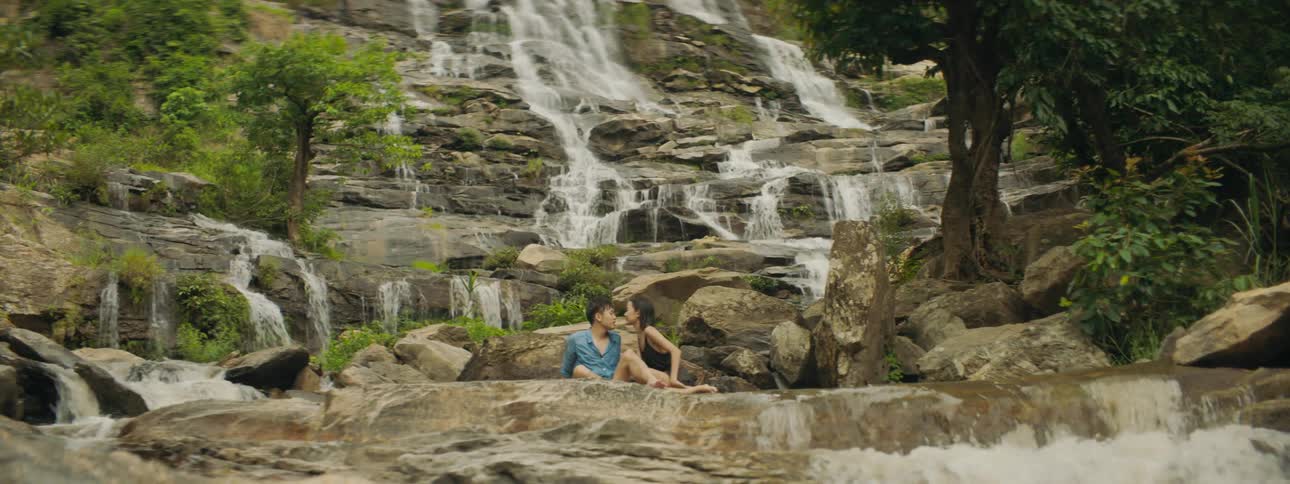 A woman kisses a man as they sit together at the bottom of a waterfall in the wilderness during the day