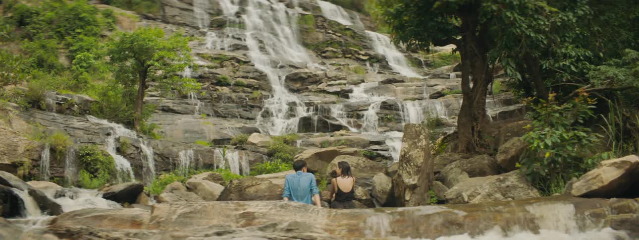 A man and woman in drenched clothing sit on a stone formation at the base of a waterfall in the wilderness during the day