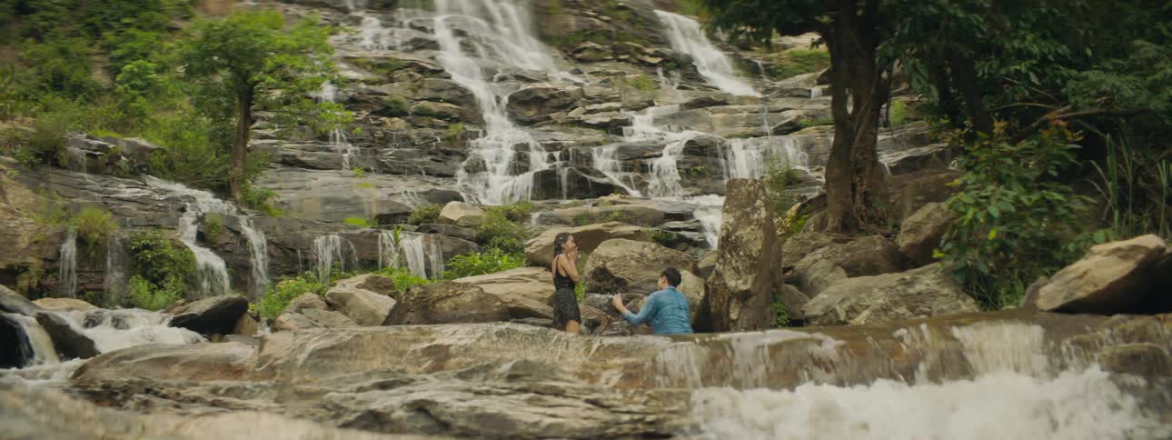 A male and female splash each other from a waterfall alongside a mountain cliff