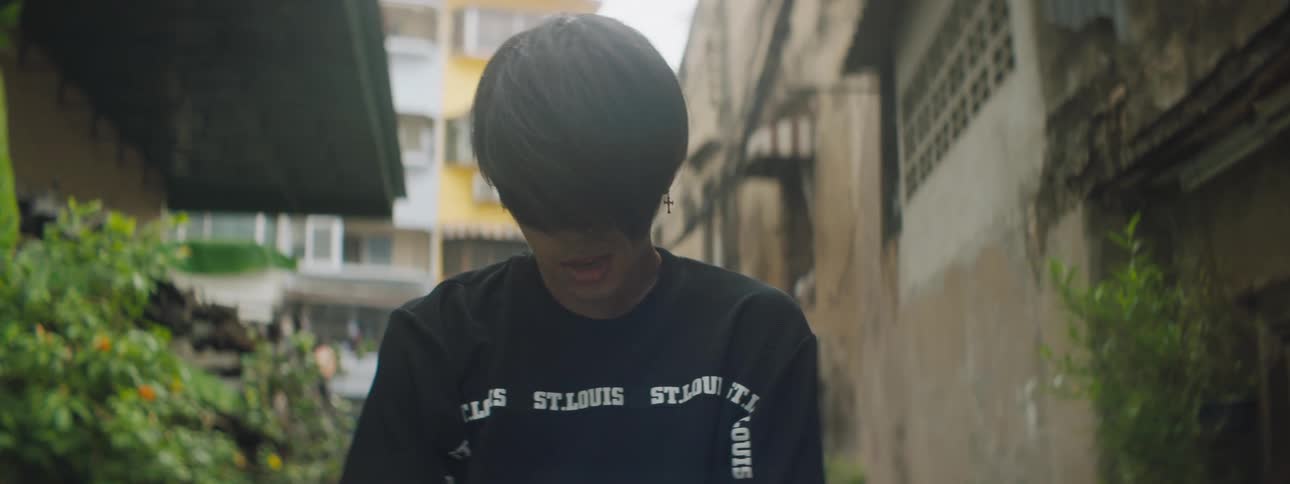 A young man poses with his head down and his hands clasps in an alleyway of a city during the day