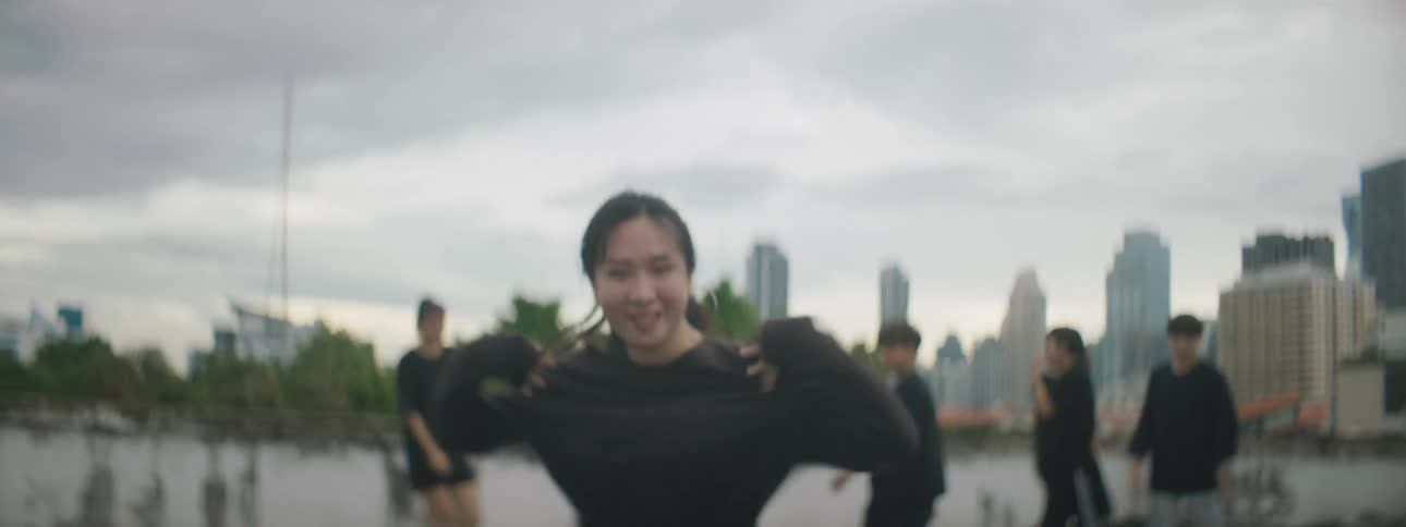 A young woman looks into the camera as she performs a pop dance on a rooftop in a metropolis on a cloudy day while a group of dancers wait behind her for their turn