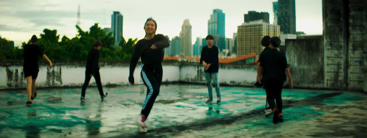 Dance troupe performs routine on roof backed by city skyline and vegetation