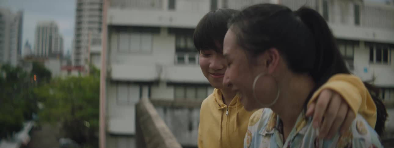 A woman sits with her arm around another woman in a neighborhood and they laugh