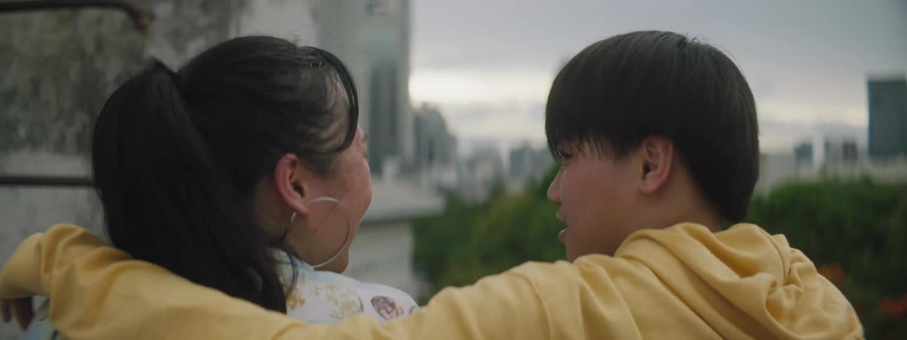 A young man kisses a young woman on the cheek as they sit together outdoors in a metropolis on a cloudy day