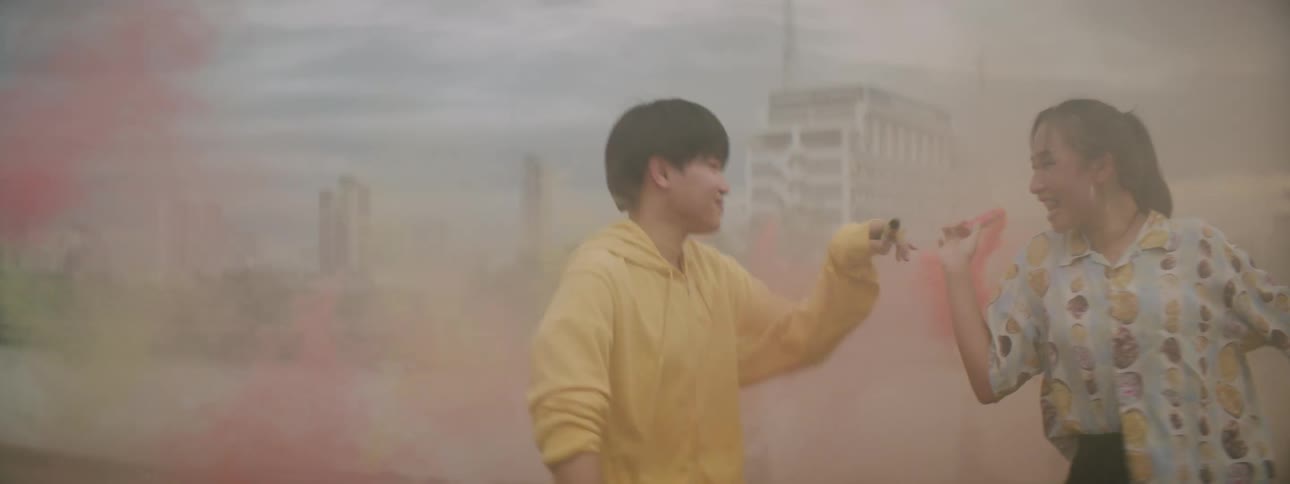 A young asian man and woman dance on a rooftop within the colorful smoke being emitted by the smoke bombs in their hands