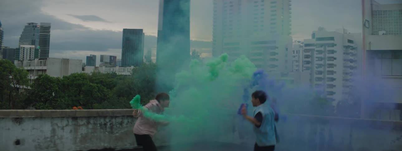 Two boys one in blue and one in orange dance on a roof top with blue and green smoke at sunset with a city skyline and large buildings behind them