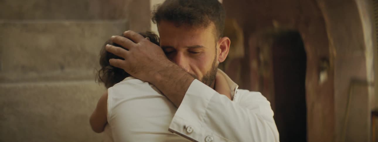 A middle eastern man standing in a stone hallway and wearing white is holding and comforting an infant baby in a white shirt