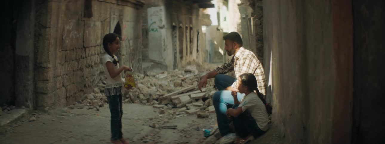 A girl shares her potato chips with her sister who sits next to their father in a alleyway between ruined buildings