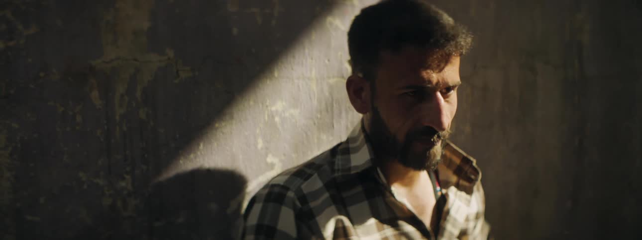 Man with a beard and a shirt leans against a wall in the sunlight