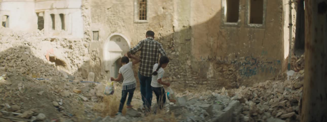 A man holds hands and walks with two little girls through rubble and debris in a village