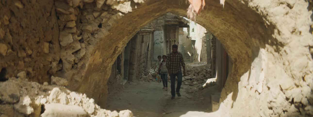 Two young girls walk behind a man through a street covered in rubble