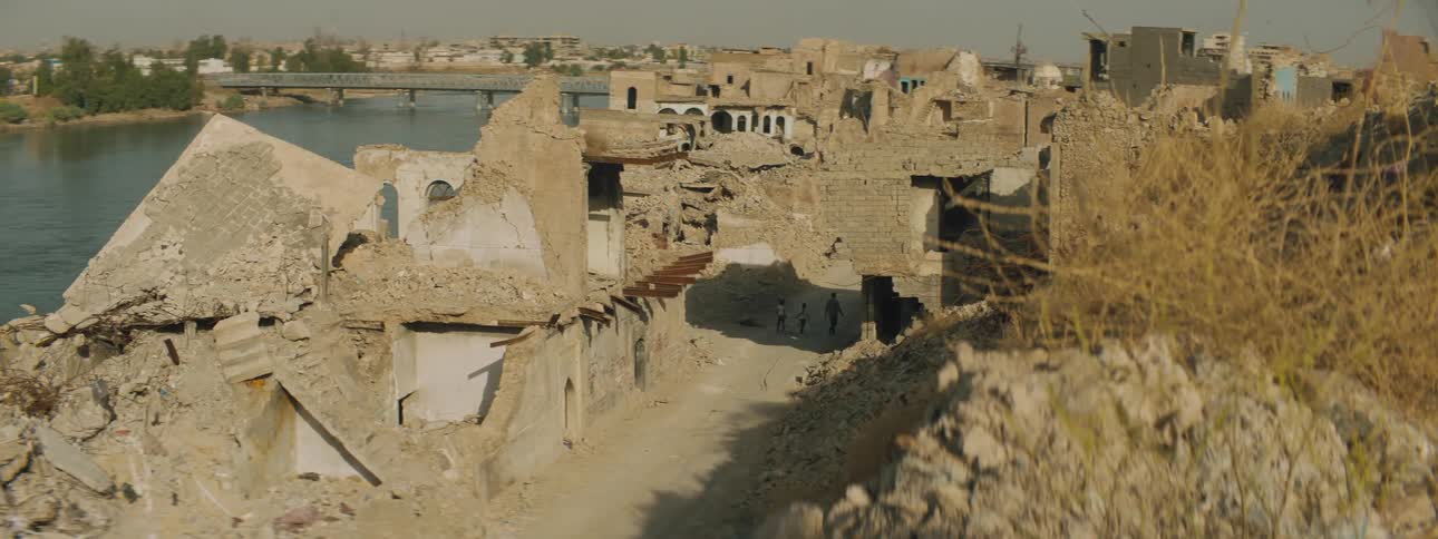 Man and girls walk along road through ruined neighborhood across river from rest of town under gray sky