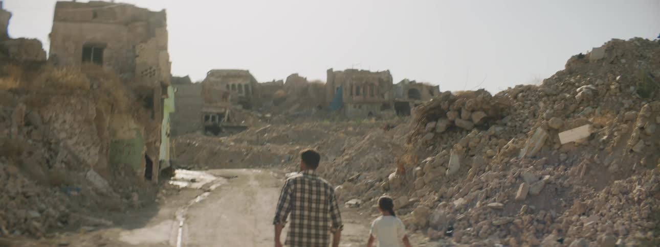 A man walking with two girls holding bags with debris dilapidated buildings the sky and clouds in the background