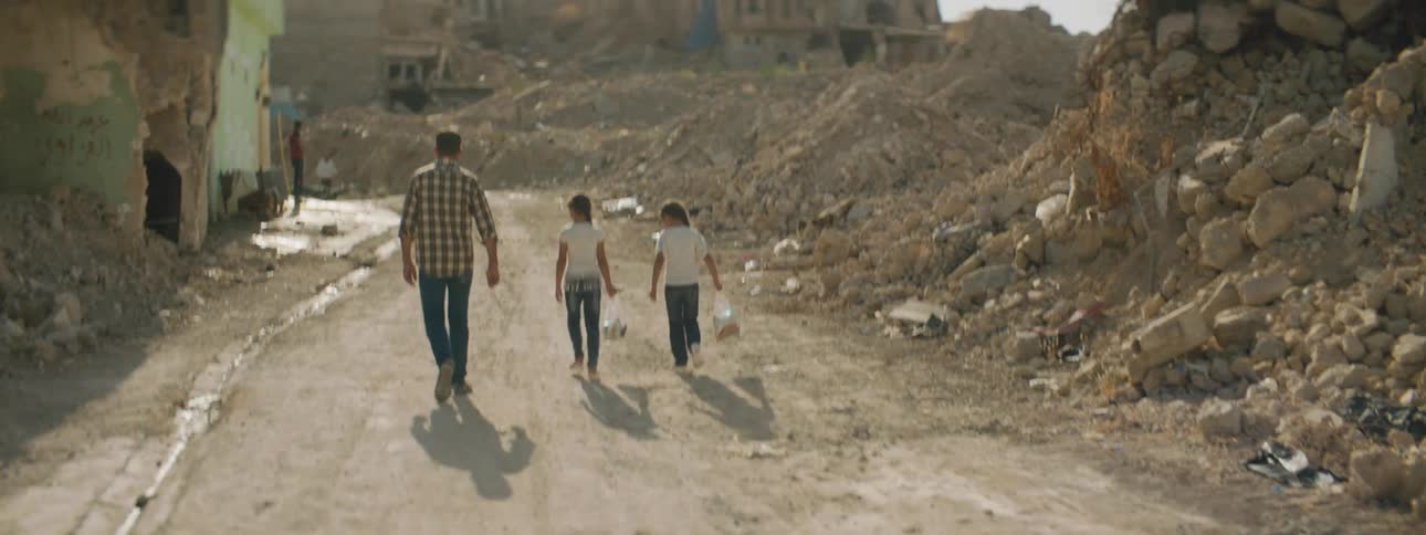 A man walks with two boys carrying bags down a dirt road with structures built into the hillside in the background