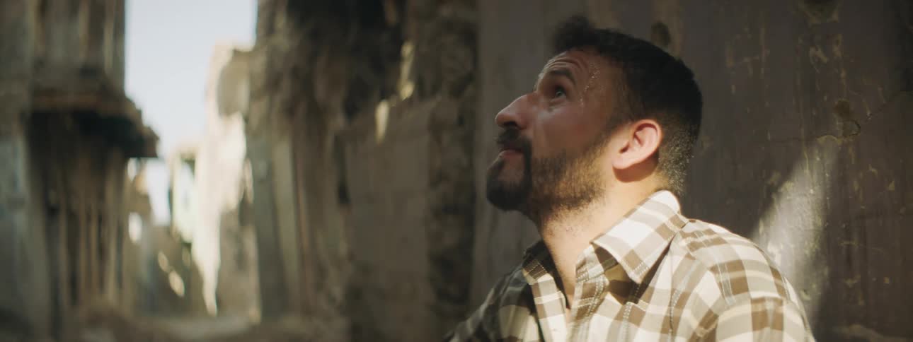 A man sitting outdoors in a ruined city looks around at the derelict buildings around him
