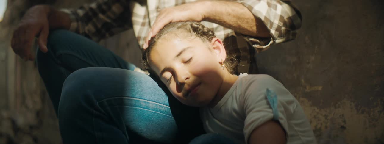 A young girl rests her head on her fathers leg as he strokes her hair outdoors on a sunny day