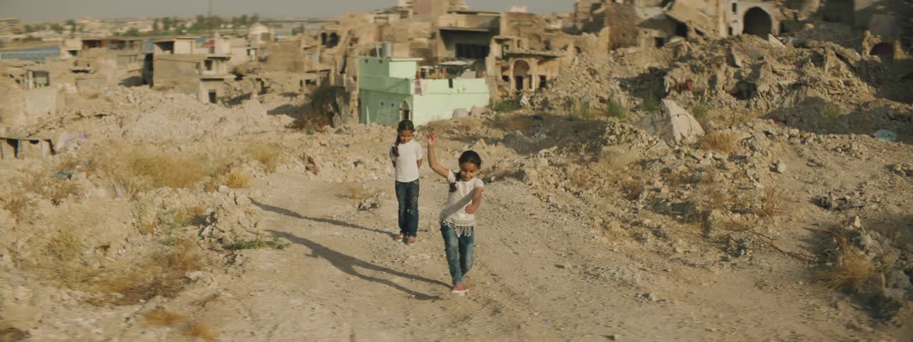 Two girls play hopscotch on soil in front of a run down neighborhood