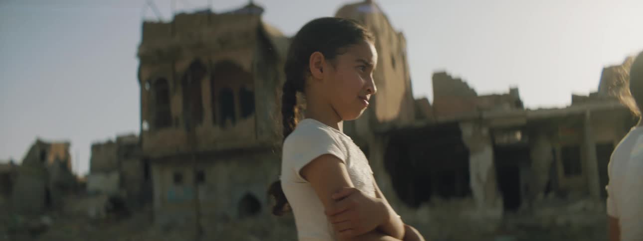 A girl stands with her arms crossed outside of a ruined building next to another girl