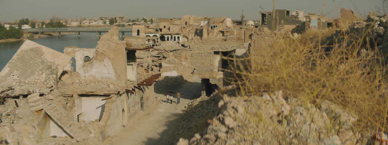 Man and two children walk along dirt road between damaged buildings across river from rest of town