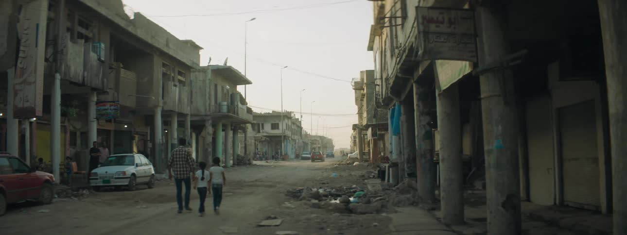 An adult and two children walk on a dirt road beside deserted buildings in a small town