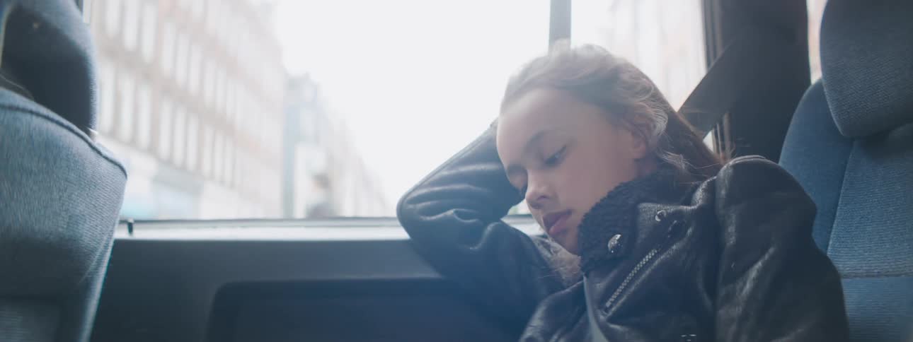 A sleepy white girl with a black jacket rests her head on a car window as she rides through the city