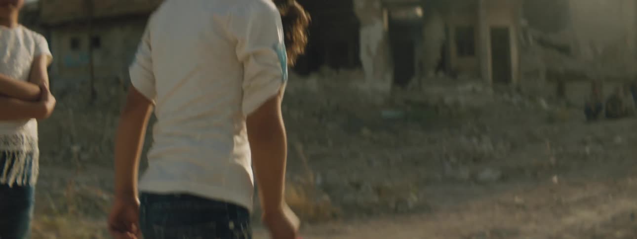 A girl plays outdoors in front of the ruins of a building as another girl stands behind her with her arms crossed