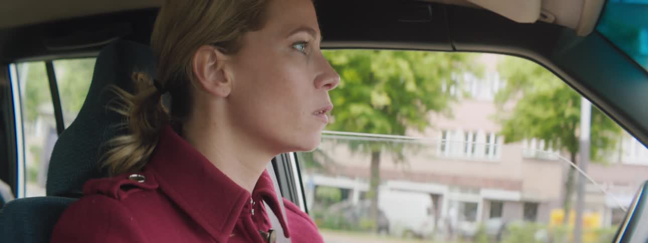 Woman drives past buildings and storefronts wearing reddish clothing