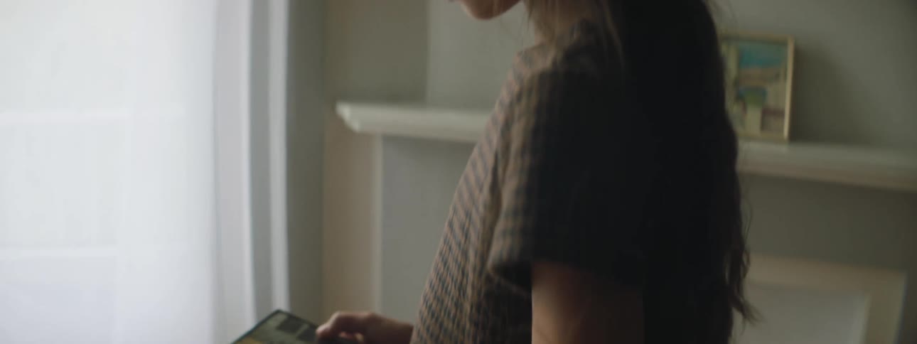 A girl gazes somberly at a photograph of a man while standing inside of a house during the day