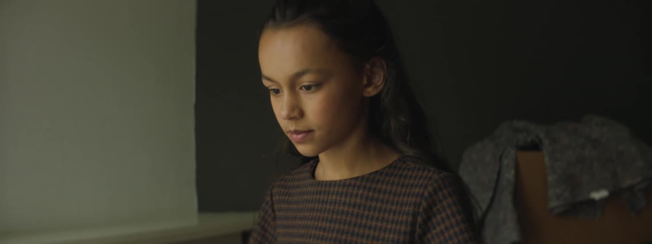 Girl with dark brown hair and a sweater looks down in a room