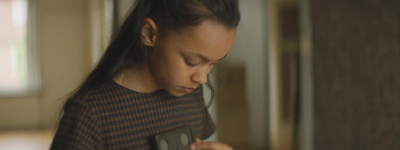 A girl standing in a room looking at a picture frame and rubbing it in her shirt with a doorway and a window in the background