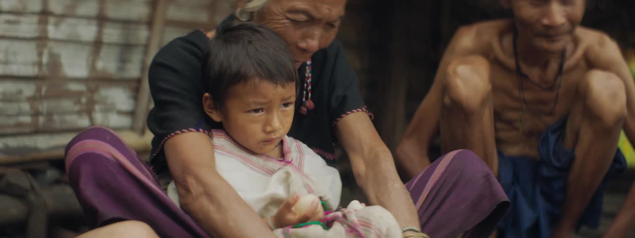 Grandparents sit outside near wooden wall smiling while grandmother holds grandchild