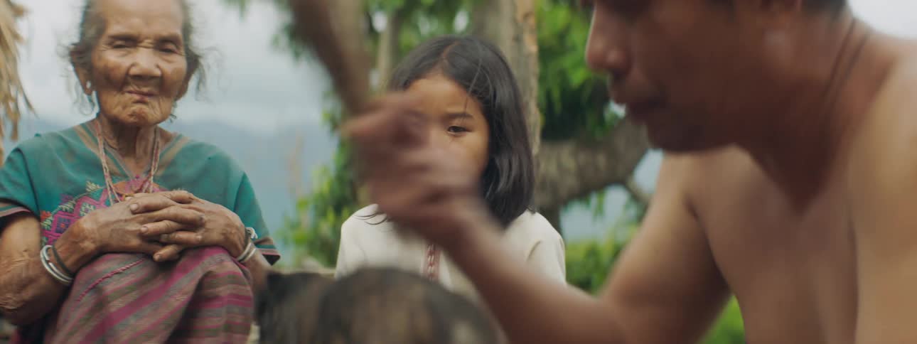 An elderly woman and a child watch as a man hits a bird with a stick to kill it
