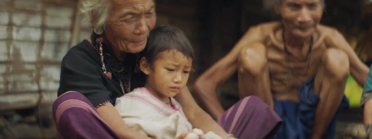 A woman in a third world village holds her grandson in her lap as she sits with others in a shelter