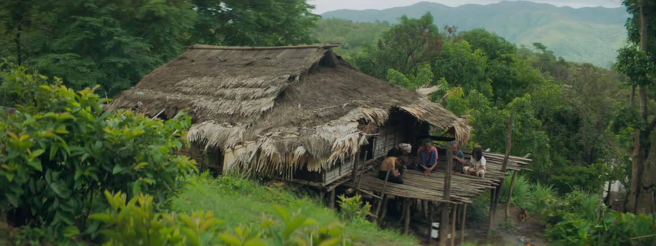 Family sits on second story of thatched hut at end of dirt path nestled against forest and hill