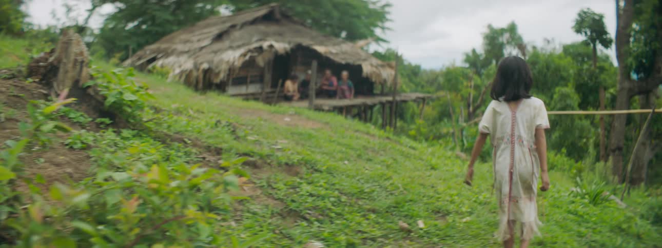 Girl walks down dirt path along slope toward family sitting on balcony of thatched hut