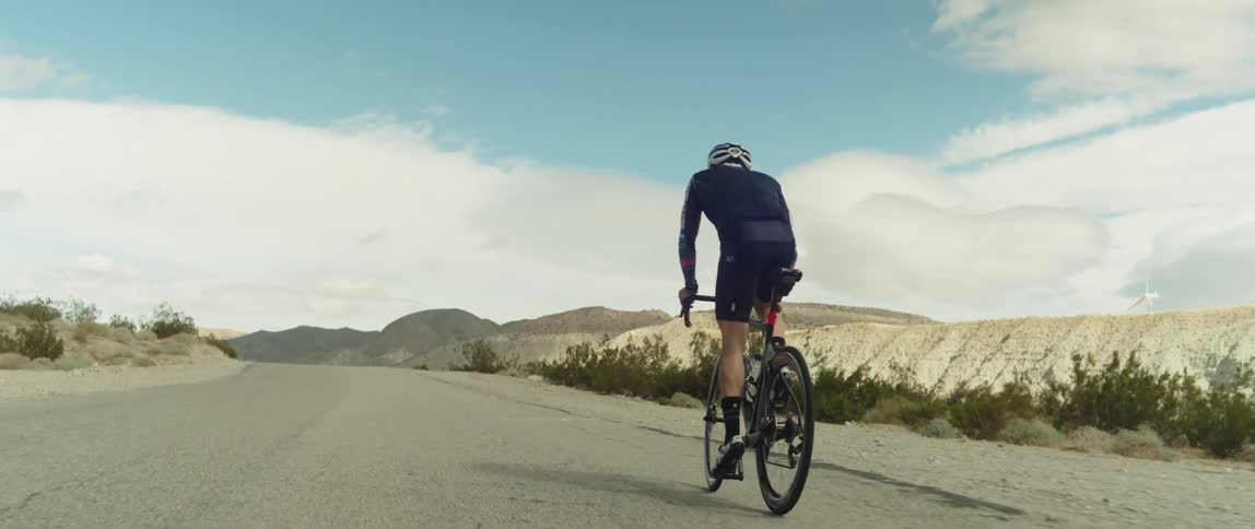 A man wearing a bicycle helmet is standing as he pedals his bike on a road near mountains