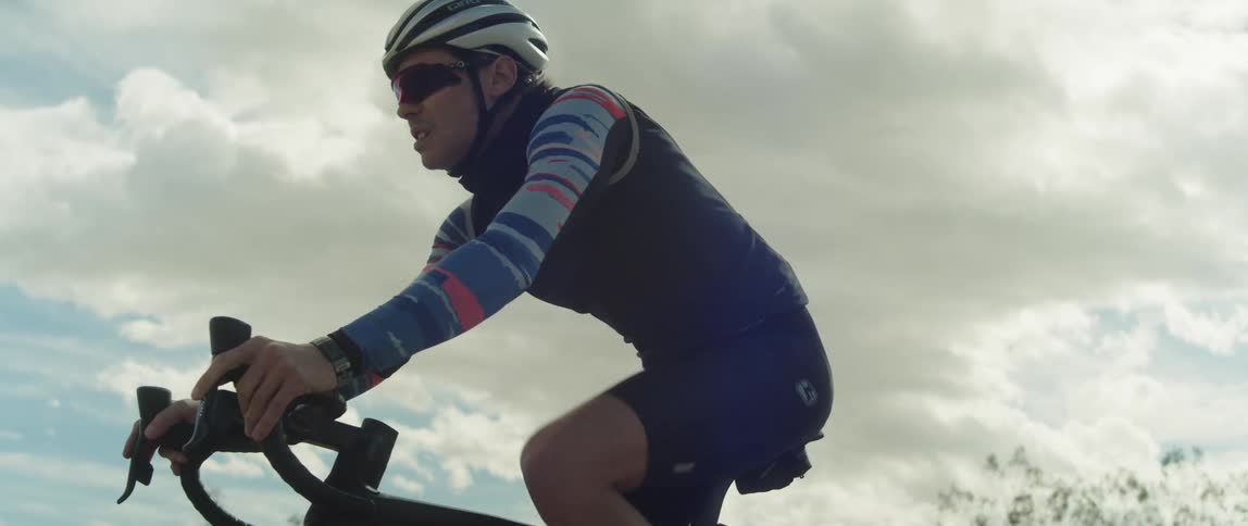 A man wearing a helmet and sunglasses is riding a bike under a cloudy sky