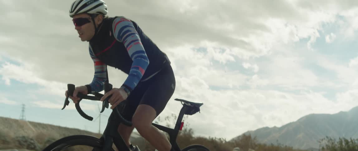 A man wearing a helmet and sunglasses is riding a bike under a cloudy sky and there are mountains in the distance behind him