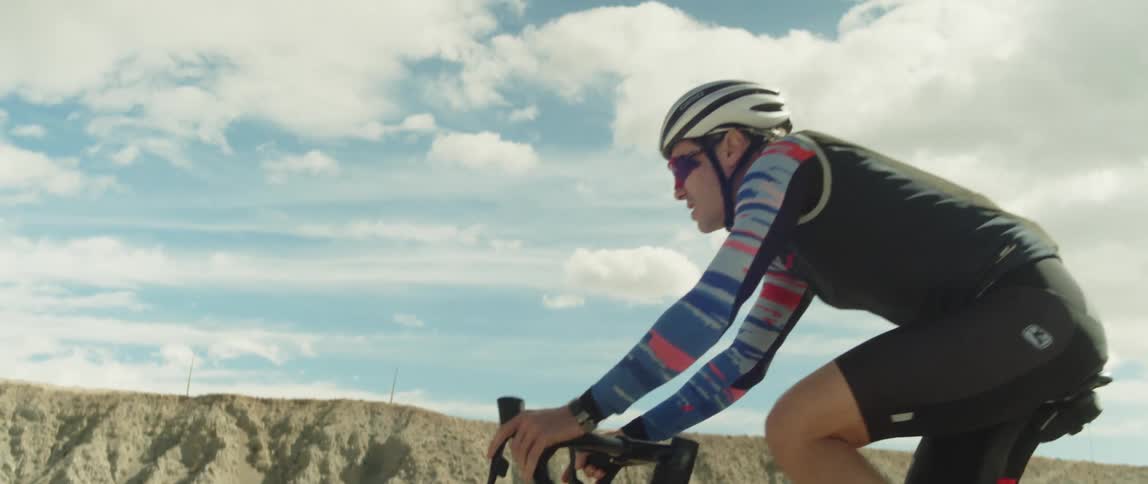 A man wearing a helmet and sunglasses is riding a bike under a cloudy sky