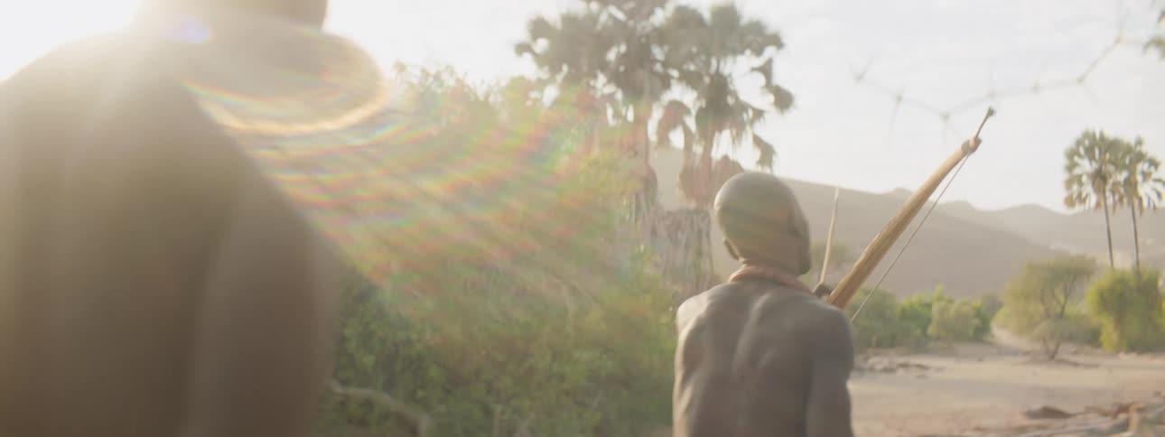 Man holding bow with arrow leads others past brush as sun shines down over mountain