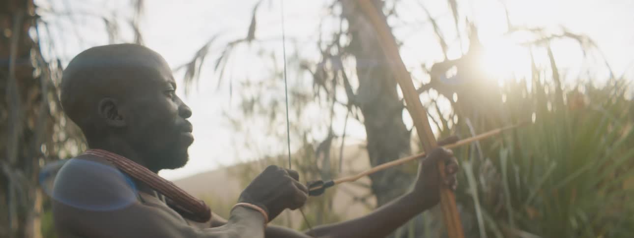 A man shooting a bow and arrow with trees the sky and clouds in the background