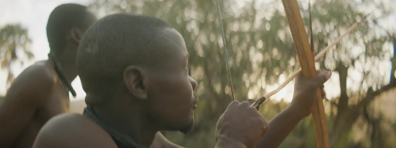 Men stalk wilderness as sun shines over mountains with drawn bow and arrow